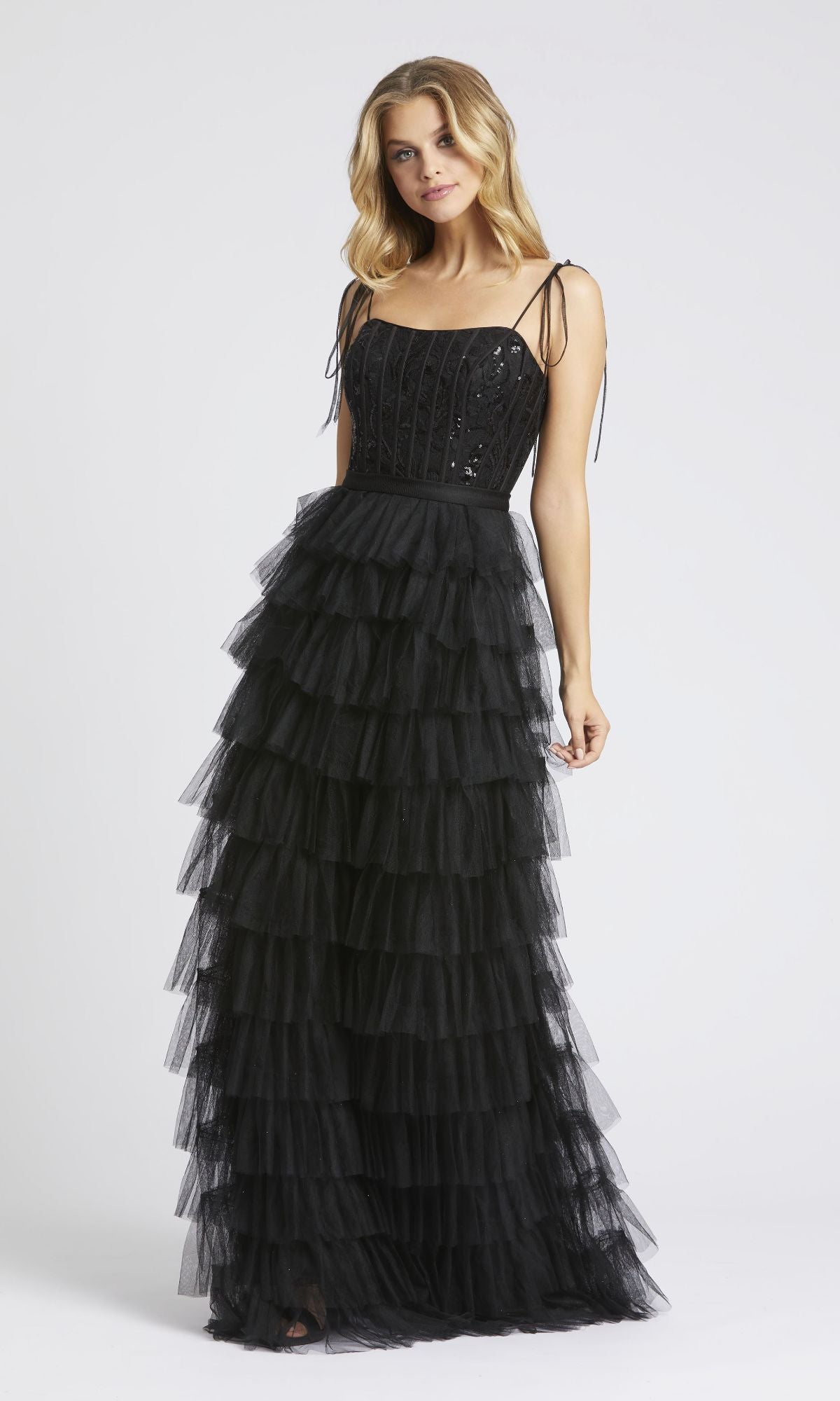 A woman models the Mac Duggal 26281, a long black ruffled prom dress with a sequined fitted bodice and tiered tulle skirt, standing against a plain white background.